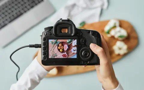 Dłoń trzyma aparat fotograficzny, na którego ekranie widać zdjęcie jedzenia: pomidorki, figi, pestki i liście.