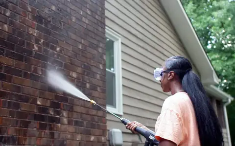Woman pressure washing a dark acid wash brick wall, restoring its natural beauty.