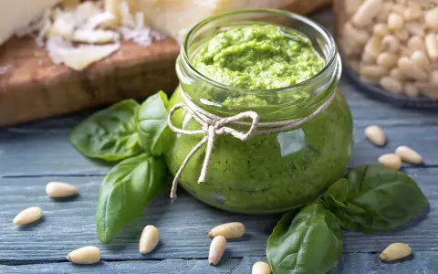 Słoik domowego pesto z bazylii, otoczony świeżymi liśćmi bazylii i orzeszkami pinii. Obok kawałki parmezanu. Idealne do makaronu.