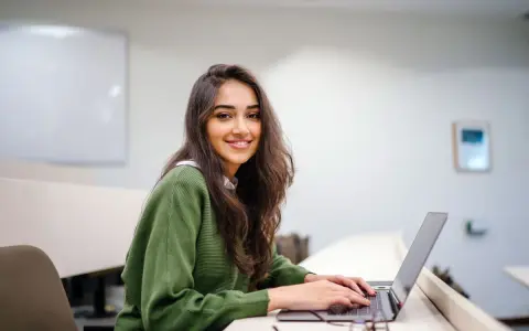 Uśmiechnięta studentka pracuje na laptopie w sali wykładowej. Jej status ucznia jest widoczny w otoczeniu i skupieniu na nauce.