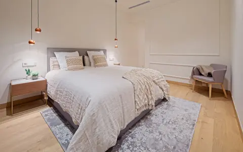 Dormitorios relajantes con cama amplia, manta texturizada, sillón acogedor y luces colgantes.
