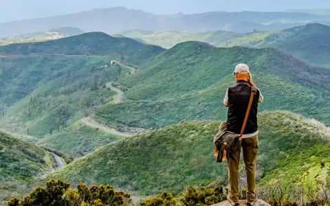 Mann mit Kamera genießt die Aussicht auf kurvige Straße in grünen Bergen. Perfektes Ziel für Wandern in Spanien.
