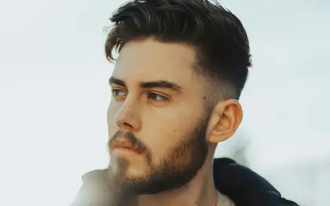 Ein Mann mit Bart und einem modernen taper fade design im Haar blickt nachdenklich zur Seite.