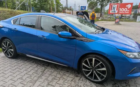 Błękitny Chevrolet Volt z czarnym dachem, zaparkowany na mokrej kostce brukowej. W tle widać stację KFC.