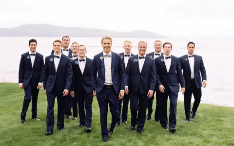 Groomsmen in navy suits with bow ties walk on grass by a lake. Can you wear a normal suit to a black tie event? This image shows a stylish alternative.