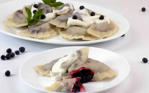 Pyszne pierogi z jagodami, polane śmietaną i ozdobione miętą. Na talerzu widać też kilka świeżych jagód.