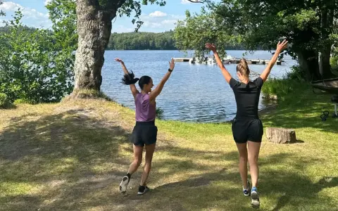 Två kvinnor hoppar glatt vid en sjö, redo att springa en mil om dagen i den vackra svenska naturen.