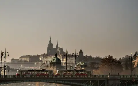 Most w Czechach, po którym przejeżdża tramwaj, z widokiem na zabytkowe budowle w oddali.