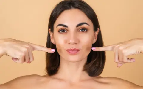 Doigts pointés vers les joues, une femme montre comment repulper son visage pour un look plus jeune et plein.