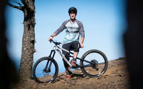 Sam Pilgrim, bereit für die nächste Abfahrt, posiert mit seinem Mountainbike auf einem felsigen Hang vor blauem Himmel.