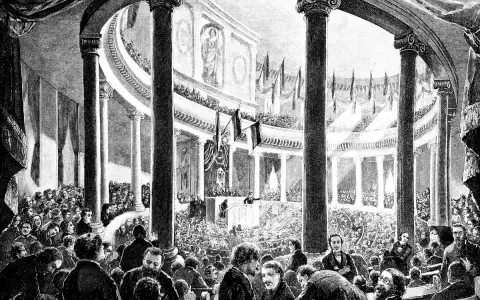 Versammlung der Frankfurter Nationalversammlung in der Paulskirche. Viele Männer in Anzügen und Hemden sitzen und stehen in einem großen, säulenreichen Gebäude.