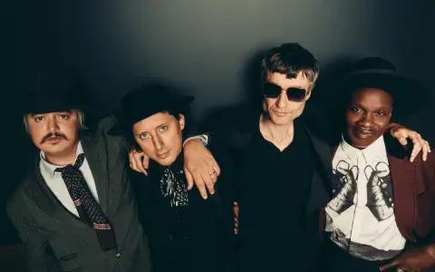 Quatre hommes stylés, comme des libertins modernes, posent ensemble. L'un porte un chapeau et une veste rayée, un autre un chapeau noir et une chemise à jabot. Le troisième, lunettes de soleil, est en noir. Le dernier, chapeau et veste bordeaux, arbore...