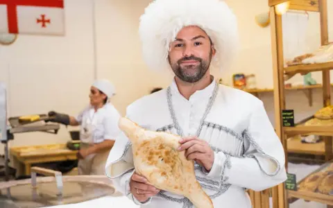 Mężczyzna w tradycyjnym stroju i czapce z owczej wełny trzyma chleb gruziński. W tle widać flagę Gruzji i piekarnię.