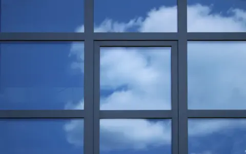Szpros dzieli okno na kwadratowe pola, w których odbija się błękitne niebo z chmurami.