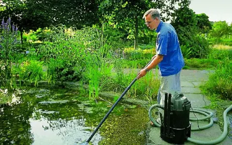 Mężczyzna wykonuje czyszczenie oczka wodnego za pomocą specjalnego odkurzacza. Wokół bujna zieleń.