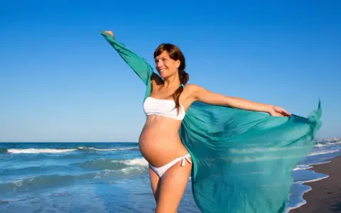 Donna incinta in bikini bianco sulla spiaggia, con un velo turchese che svolazza al vento. Il mare sullo sfondo.