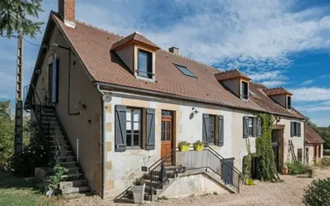 Une maison de campagne simple avec des volets gris, une cheminée et des escaliers extérieurs sous un ciel bleu.