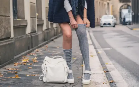 Stylizacja na jesień: jak nosić długie skarpetki damskie do sukienki i butów na płaskiej podeszwie. Biały plecak i liście na chodniku.