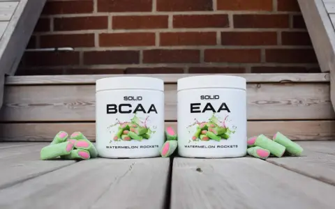 Två burkar med kosttillskott, en märkt BCAA och en EAA, står framför en tegelvägg. Runt burkarna ligger gröna godisar med rosa inuti, som ser ut som vattenmelonraketer.
