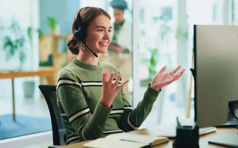 Uśmiechnięta customer experience manager w słuchawkach gestykuluje, siedząc przy biurku przed monitorem.