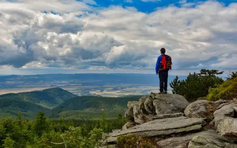 Turysta z plecakiem podziwia rozległą panoramę Gór Opawskich. Chmury na niebie tworzą dramatyczny spektakl.