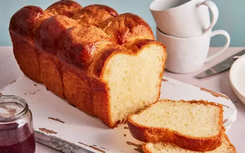Ein goldbraunes Brioche Brot Rezept, aufgeschnitten auf einem Holzbrett mit Marmelade und Tassen im Hintergrund.