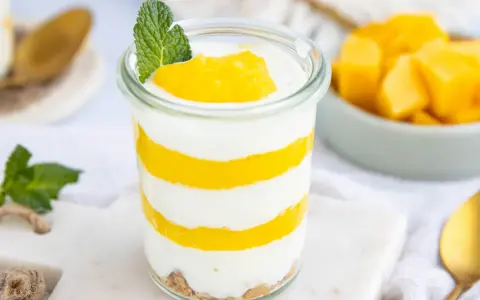 Schichtweise geschichtetes Mango-Parfait in einem Glas, garniert mit Minze. Daneben eine Schüssel mit frischen Mangostücken.