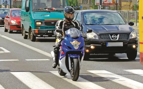 Motocyklista w kasku i kombinezonie na pasach. Obok samochody i furgonetka. Pamiętaj o przeglądzie, bo jaka kara za brak przeglądu może być dotkliwa.