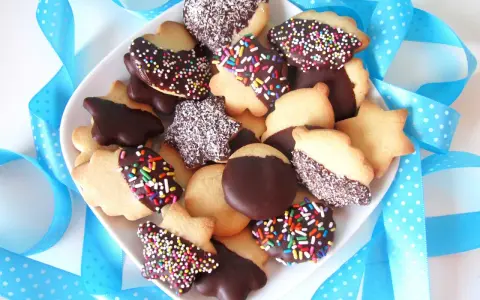 Un plato lleno de pastas de té caseras, algunas cubiertas de chocolate y chispas de colores, otras con coco rallado, rodeadas de cintas azules.