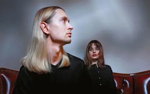 Łukasz Rozmysłowski z długimi blond włosami i kobieta z brązowymi włosami siedzą na skórzanej kanapie.