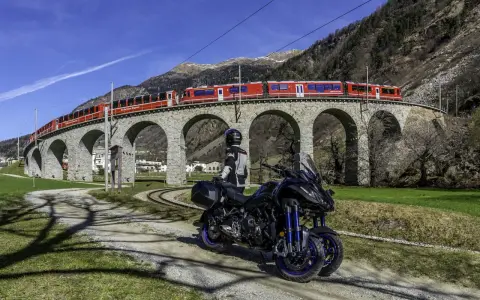 Motociclista e moto su strada sterrata, con un treno rosso che attraversa un viadotto ad archi in pietra. Paesaggio alpino con montagne sullo sfondo, vicino al passo della Novena.