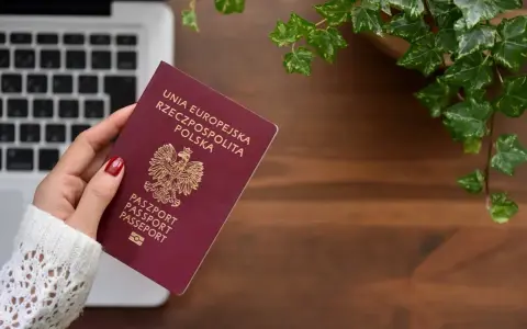 Dłoń trzyma polski paszport na tle laptopa i roślin.