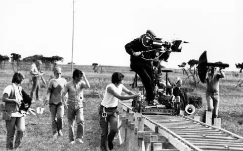 Sul set di un film, un operatore manovra una cinepresa su binari per un piano sequenza. Altri membri della troupe osservano e lavorano.