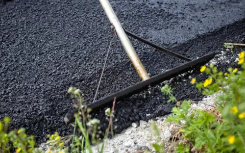 Verktyg sprider ny asfalt för att asfaltera uppfart. Grönska och gula blommor växer vid sidan.