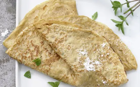 Drei goldbraune Dinkel Pfannkuchen, leicht mit Puderzucker bestäubt und mit frischer Minze garniert, auf einem weißen Teller.