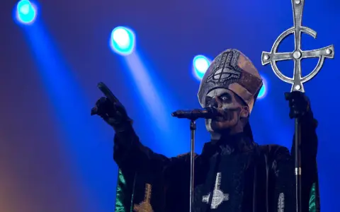 Jocke Berg som Papa Emeritus IV från Ghost, med vit ansiktsmålning och en korsstav, pekar mot publiken under en konsert.