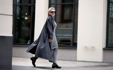 Elegante Frau ab 70 mit Kurzhaarschnitt und Sonnenbrille, die in einem langen grauen Mantel und glitzerndem Kleid durch die Stadt geht.