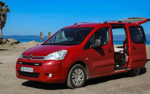Czerwony Citroen Berlingo 1.6 HDI na plaży, z otwartymi drzwiami i bagażnikiem, na tle morza i palmy.
