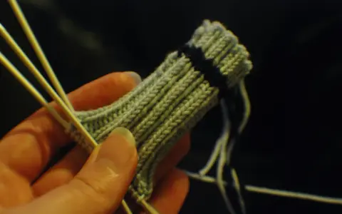 En hand håller stickor och ett ljusgrönt stickat tyg med en mörk rand. Det ser ut som att någon ska sticka avig maska.