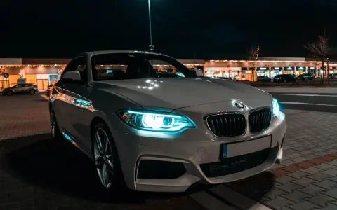 Białe BMW z włączonymi światłami adaptacyjnymi błyszczy nocą przed sklepami.