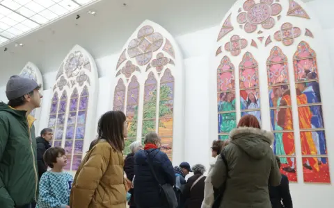 Des visiteurs contemplent de grands vitraux contemporains aux couleurs vives, représentant des scènes et des paysages.