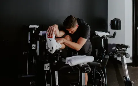 Man in black shirt stretches his outer hamstring by holding his foot, surrounded by stationary bikes in a gym.