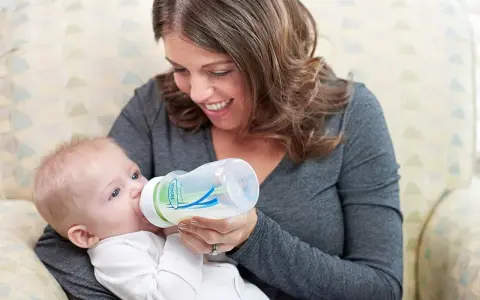 Uśmiechnięta mama karmi niemowlę butelką z systemem odpowietrzania. Smoczek do butelki Dr Brown zapewnia komfortowe karmienie.