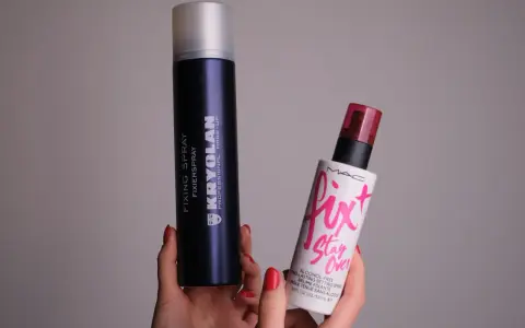 Dwie ręce trzymają dwa spraye: duży, granatowy Kryolan Fixing Spray i mniejszy, biały Fix+ Stay Over MAC. Oba to profesjonalne produkty typu fixer.