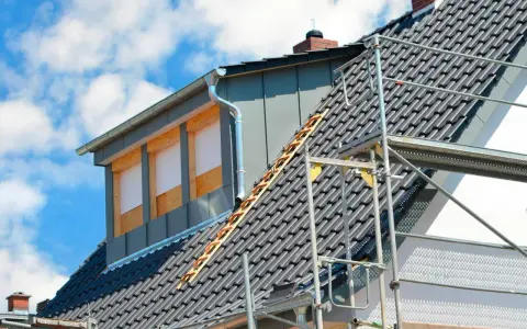 Bygga takkupa pågår. Ett hus med takpannor och byggställning, en ny takkupa med fönsteröppningar syns.