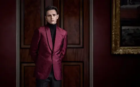 A man in a patterned burgundy jacket and black turtleneck, perfect for cocktail party attire for men, stands in a dimly lit room with ornate wooden doors.