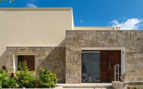Moderne facciate in pietra grezza si alternano a intonaco chiaro, creando un contrasto materico. Un ingresso con porta in legno scuro e un giardino fiorito.
