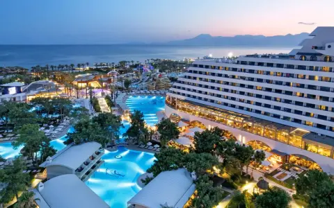 Luksusowy hotel Titanic Deluxe Lara z basenami i parkiem wodnym, oświetlony wieczorem, z widokiem na morze i góry.