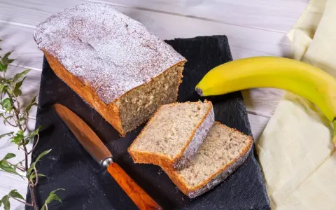 Ein **super saftiger Bananenkuchen**, bestäubt mit Puderzucker, liegt auf einem schwarzen Stein neben zwei Scheiben. Eine Banane und ein Messer liegen daneben.
