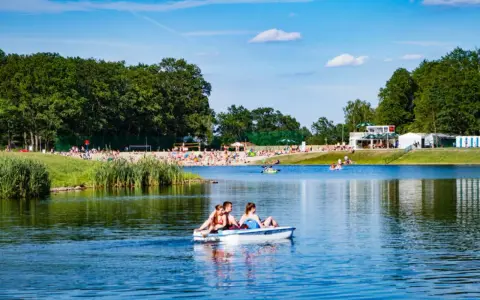 Rodzina pływa rowerkiem wodnym po jeziorze na Dolnym Śląsku, w tle plaża pełna ludzi i zielone drzewa.